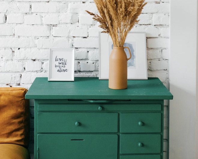 A green dresser with multiple drawers is set against a white brick wall. On top of the dresser, there is a brown vase holding dried grass, and two framed pictures—one with the text 'love will keep us alive' and another featuring an abstract artwork. Part of a mustard-colored sofa is visible on the left side.