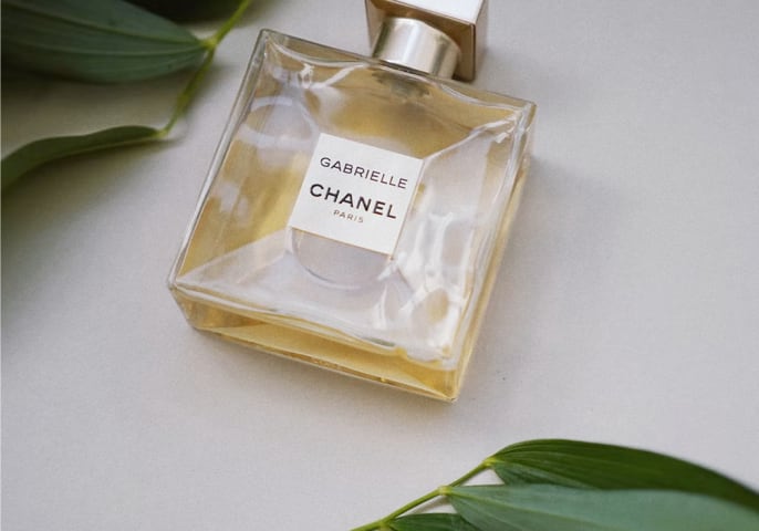 Bottle of the Gabrielle
          Essence perfume on a gray surface, surrounded by green leaves